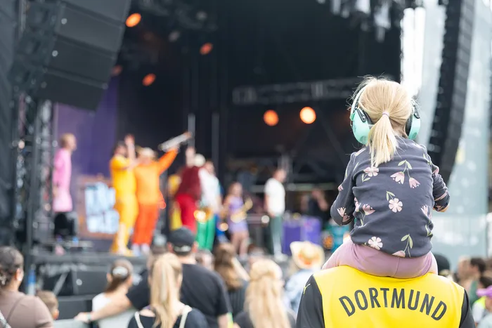 Ein Kind mit Kopfhörern sitzt auf den Schultern einer Person und beobachtet eine bunte Musikperformance auf der Bühne beim Bautz 2026.