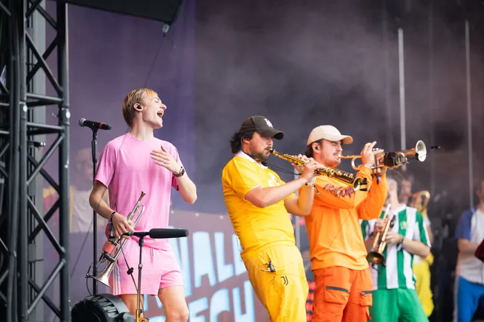 Ein Sänger in Rosa singt auf einer Bühne, während Musiker Saxophon und Trompete spielen.