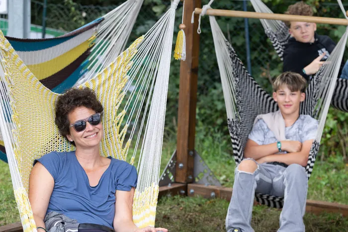 Eine lächelnde Frau mit Sonnenbrille sitzt in einer gelben Hängematte, im Hintergrund sitzt ein Junge in einer schwarz-weiß gestreiften Hängematte.