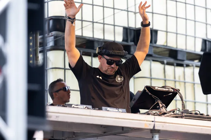 DJ Chris Newmas mit Sonnenbrille und schwarzer Kappe hebt die Arme hinter seinem DJ-Equipment.