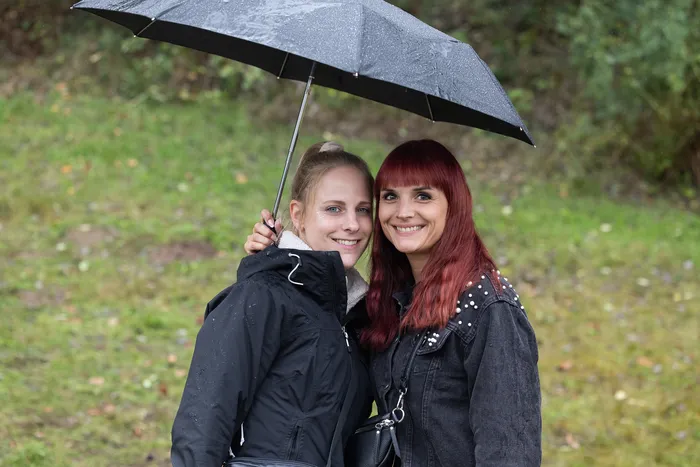 Zwei lächelnde Frauen stehen unter einem schwarzen Regenschirm vor einem grünen Hintergrund.