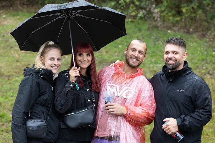 Vier lächelnde Personen stehen im Freien unter einem schwarzen Regenschirm, ein Mann trägt einen transparenten rosa Regenmantel.