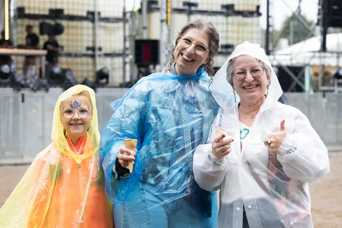 Ein Kind und zwei Frauen in bunten Regencapes lächeln vor einer Festivalbühne; eine Frau hält ein Eis, die andere ein Glas Wein.