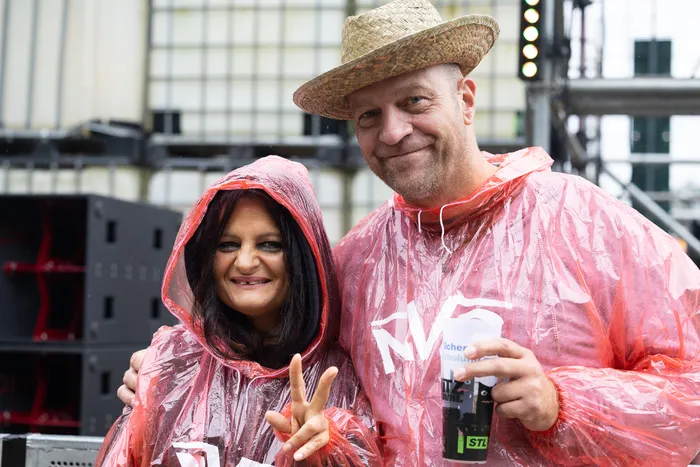 Ein Mann mit Strohhut und eine Frau in transparenten rosa Regenponchos lächeln; die Frau zeigt ein Peace-Zeichen.