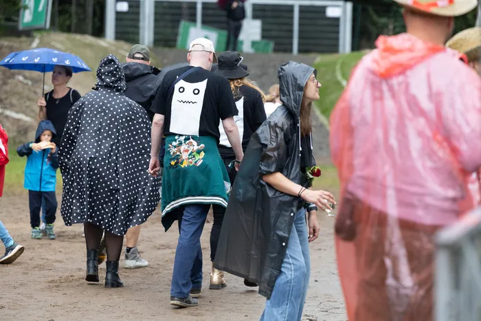 Besucher in Regenbekleidung und Ponchos gehen über einen sandigen Weg bei einem Festival.