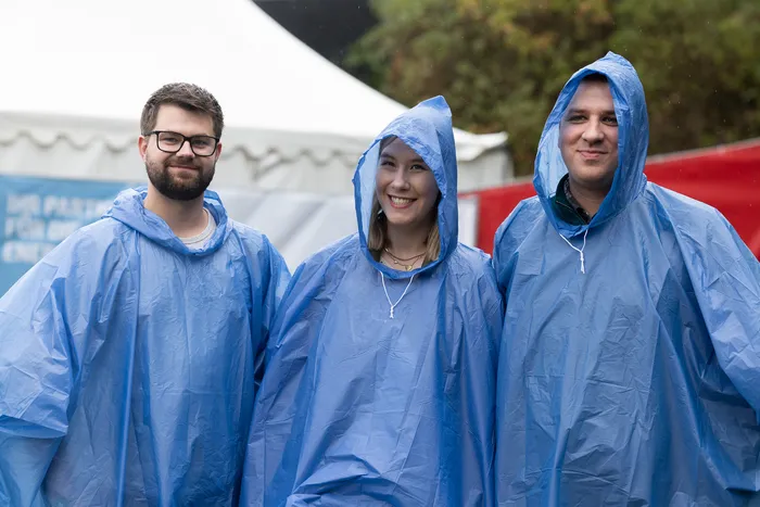 Drei lächelnde Personen in blauen Regenponchos stehen vor einem weißen Zelt auf der Bautz 2026.