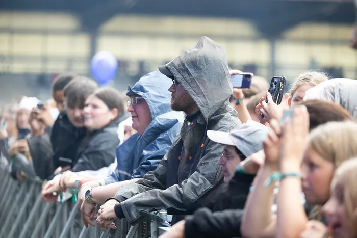 Zuschauer in Regenbekleidung stehen an einer Absperrung und halten Smartphones hoch.