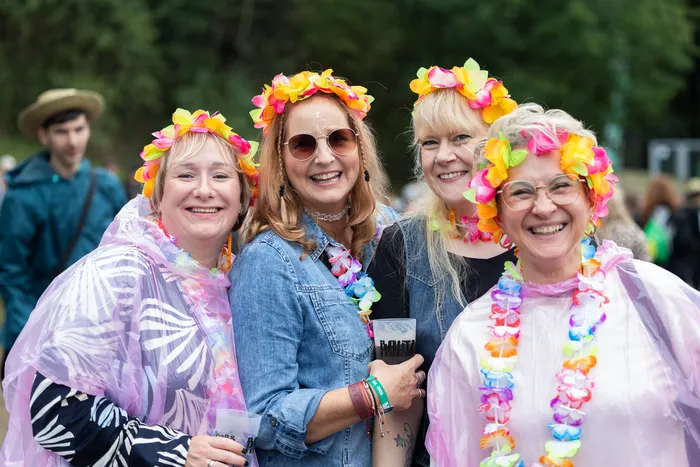 Vier lächelnde Frauen mit bunten Blumenkränzen und Blumenketten posieren gemeinsam, zwei tragen transparente Regenponchos.