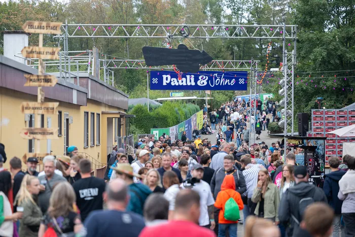 Große Menschenmenge auf einem Festivalgelände unter einem blauen Schild mit der Aufschrift „Pauli & Söhne“ und hölzernen Wegweisern.