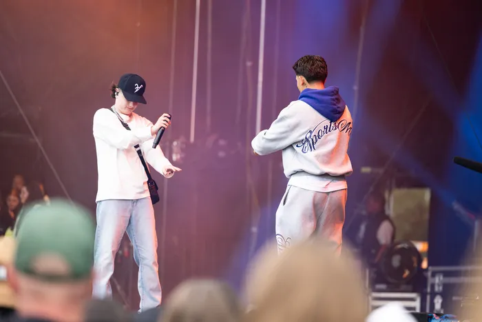 Ein Musiker in weißem Oberteil und Jeans hält ein Mikrofon auf der Bühne, während eine zweite Person in einem grauen Hoodie mit dem Rücken zur Kamera steht.