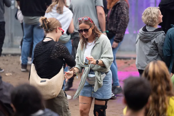 Eine Frau mit Sonnenbrille und hellem Hemd hält ein Getränk auf einem Festivalgelände.