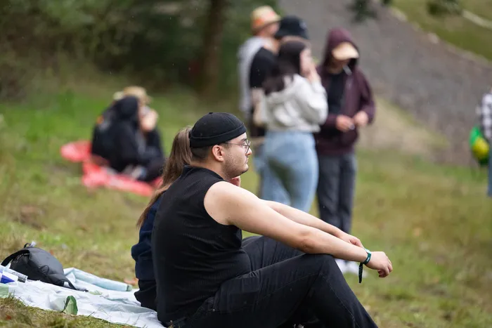 Ein Mann mit langen Haaren, schwarzer Kappe und Tanktop sitzt auf einer Decke auf einer Wiese. Im Hintergrund sind weitere Personen unscharf zu sehen.