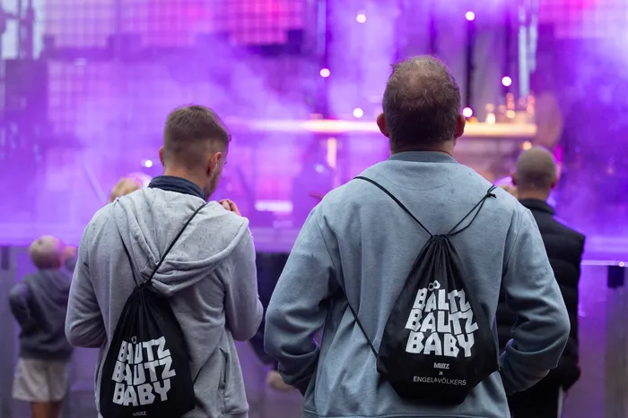 Zwei Männer von hinten, die mit schwarzen Beuteln mit der Aufschrift „BALTZ BALTZ BABY“ auf eine lila beleuchtete, vernebelte Bühne blicken.