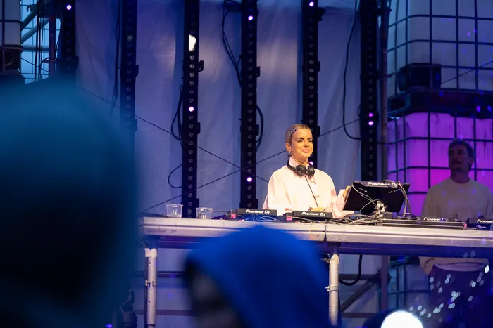 DJ Lovra steht mit Kopfhörern um den Hals hinter einem DJ-Pult mit Pioneer-Equipment auf einer blau beleuchteten Bühne.