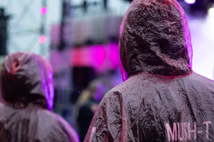 Rückansicht von Personen in dunklen Regenponchos vor einer pink beleuchteten Bühne. Auf dem Poncho im Vordergrund steht der Schriftzug „MUSH-T“.