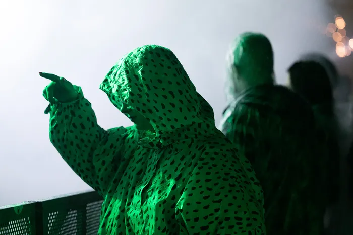 Eine Person in einem grün gepunkteten Kapuzenmantel zeigt mit ausgestrecktem Finger unter grünem Licht.