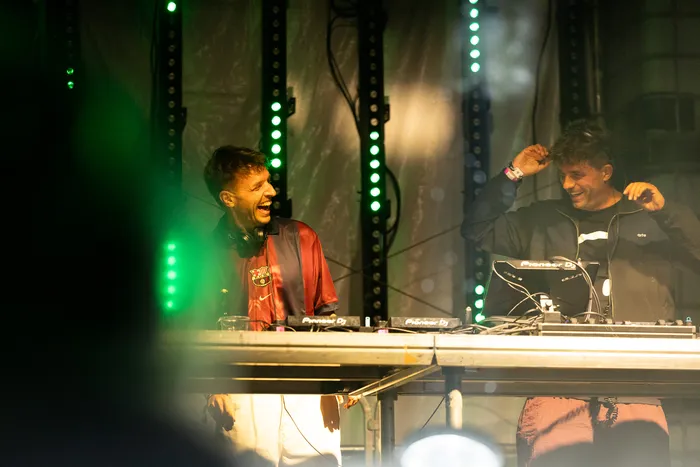 Zwei DJs hinter einem Mischpult auf einer Bühne mit vertikalen grünen Lichtstreifen; einer lacht, der andere richtet seine Kopfhörer.
