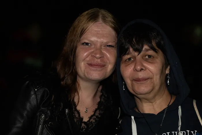 Zwei lächelnde Frauen posieren eng beieinander, eine trägt eine schwarze Lederjacke und die andere einen dunklen Kapuzenpullover.