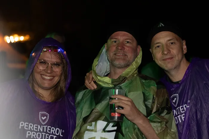 Drei lächelnde Personen in lila und grün gemusterten Regenponchos bei einem nächtlichen Event.