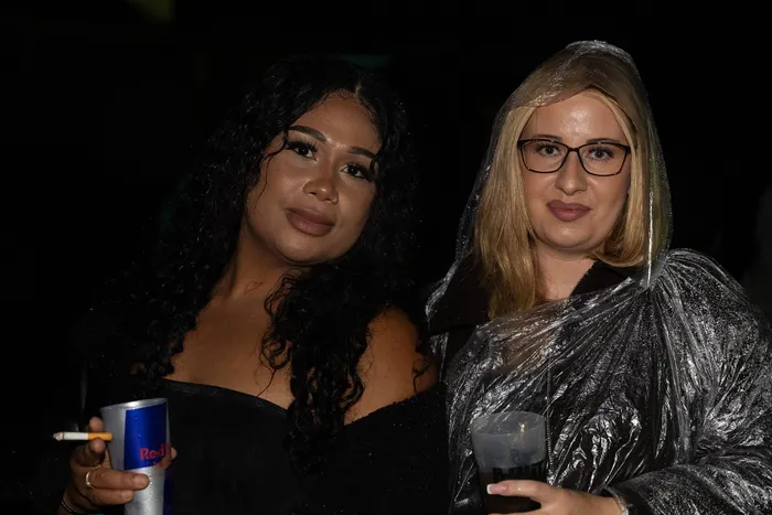 Zwei Frauen posieren nachts; eine mit dunklen Locken hält eine Dose und eine Zigarette, die andere mit Brille trägt einen transparenten Regenponcho.