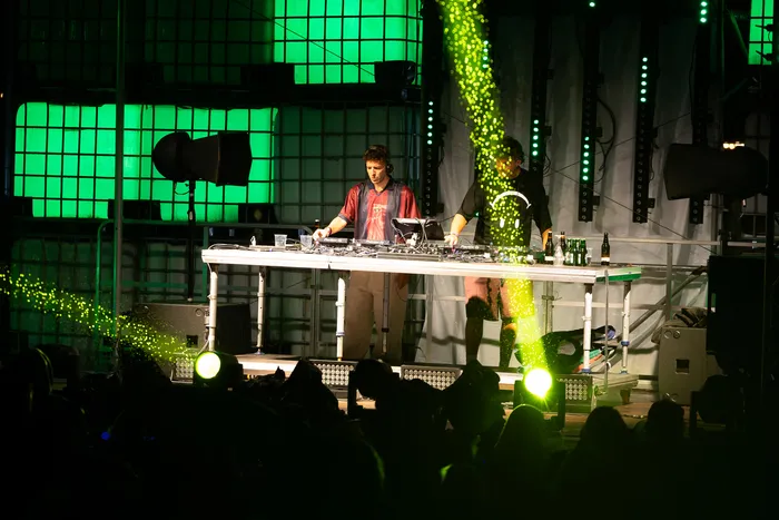 Ein DJ bedient an einem Pult mit Turntables die Musik auf einer Bühne, die von grünen Lichtstrahlen beleuchtet wird.