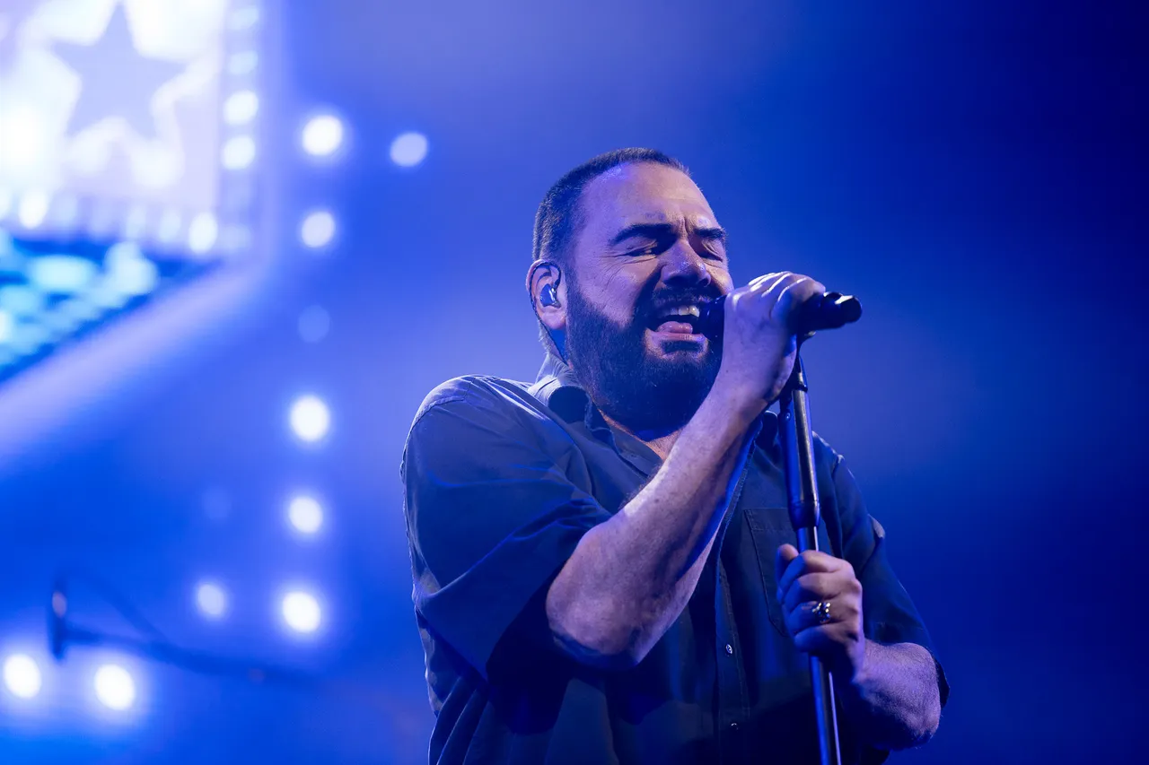 Ein bärtiger Mann singt mit geschlossenen Augen in ein Mikrofon vor blauem Bühnenlicht.