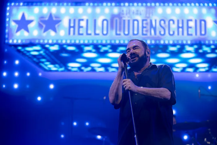 Ein Sänger singt in ein Mikrofon vor einem hellblau leuchtenden Schild mit der Aufschrift „HELLO LUDENSCHEID“.
