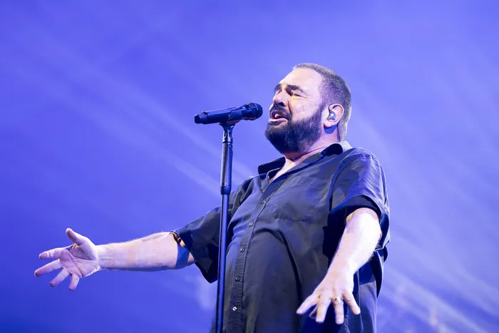 Ein bärtiger Mann in einem schwarzen Hemd singt mit ausgebreiteten Armen in ein Mikrofon vor blauem Bühnenlicht.