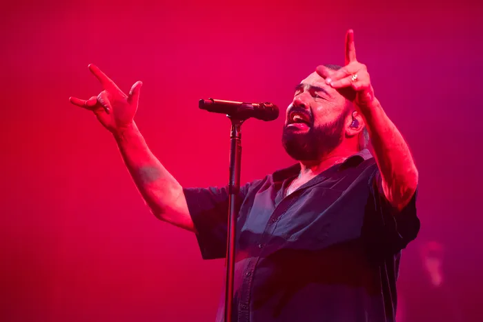 Ein bärtiger Mann singt mit erhobenen Händen in ein Mikrofon unter rotem Bühnenlicht.