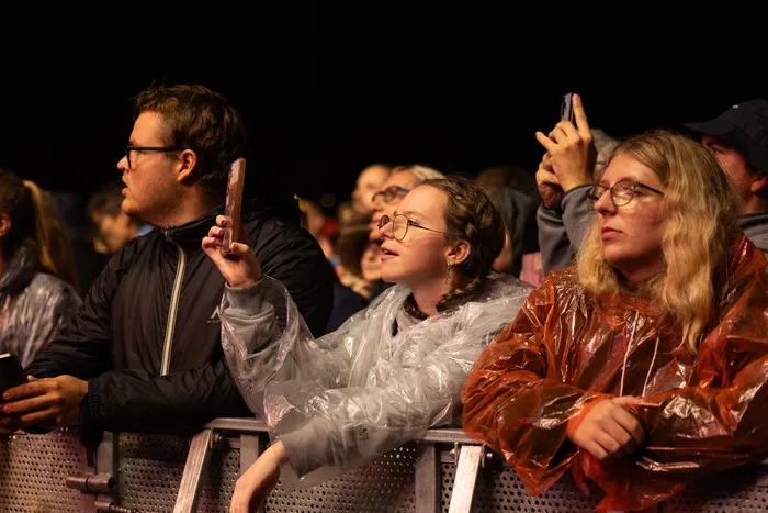 Zuschauer vor einer Absperrung bei einem Festival, darunter eine Frau in einem durchsichtigen Regenmantel, die ihr Smartphone hochhält.