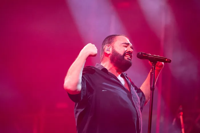 Ein bärtiger Mann singt mit geschlossenen Augen und erhobener Faust in ein Mikrofon unter roter Bühnenbeleuchtung.