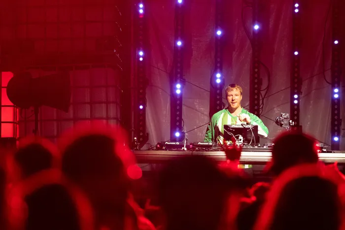 Ein DJ in grüner Jacke performt an einem Turntable auf einer rot beleuchteten Bühne vor einer Menschenmenge.