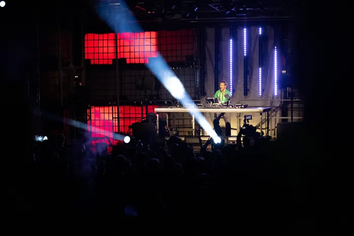 Ein DJ performt an einem Turntable auf einer Bühne mit roter und blauer Beleuchtung vor einer Menschenmenge.