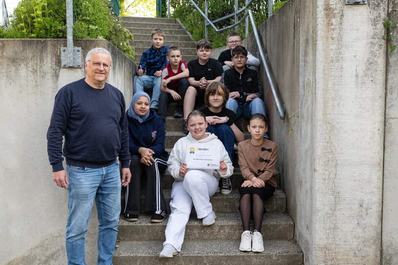 Lehrer und Schüler posieren auf einer Betontreppe, die Schülerin in der Mitte hält eine Urkunde. Im Hintergrund sind weitere Schüler zu sehen.