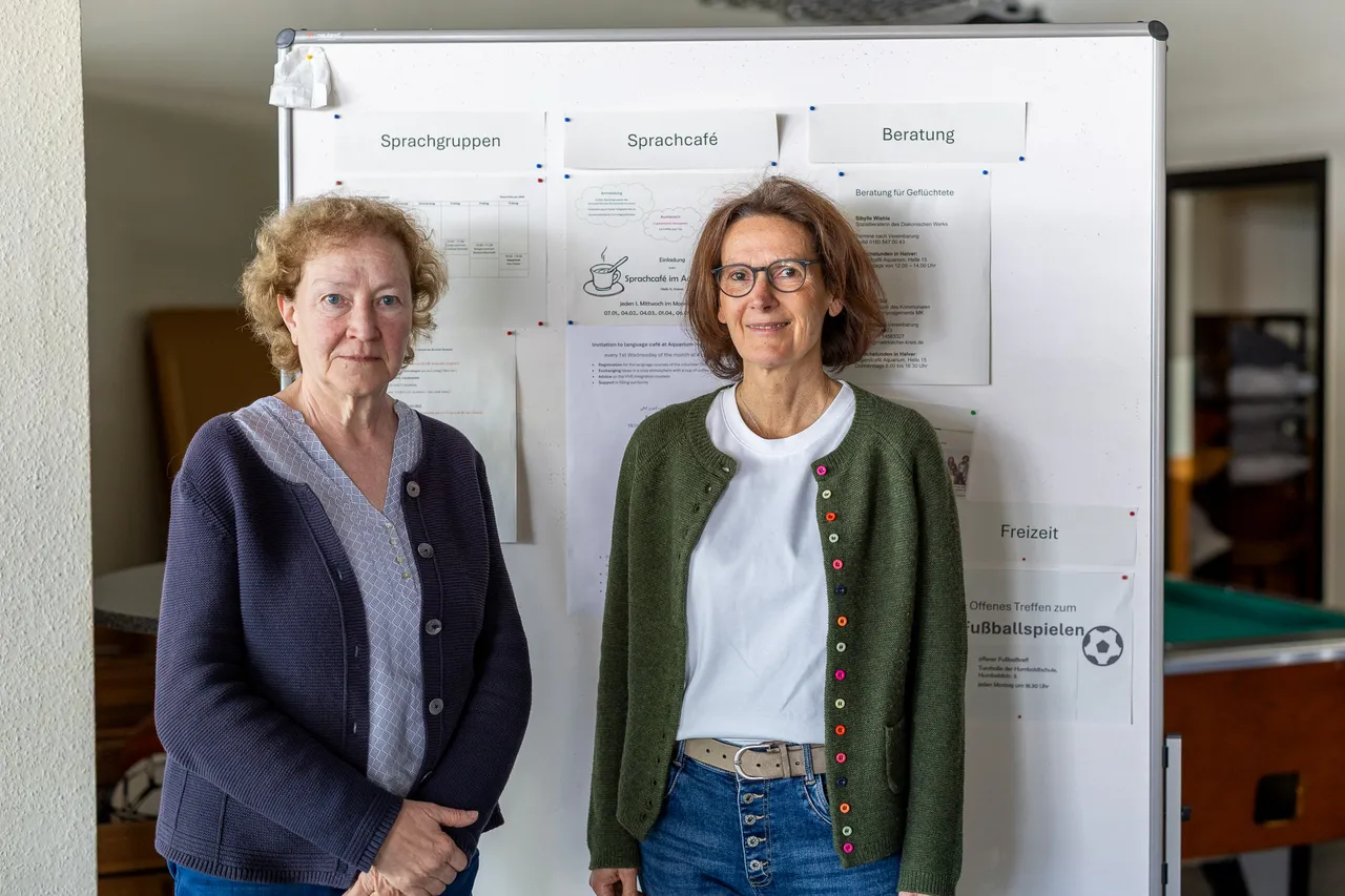 Zwei Frauen mittleren Alters stehen nebeneinander vor Plakaten mit Informationen zu Sprachgruppen, einem Sprachcafé und Beratungsangeboten.