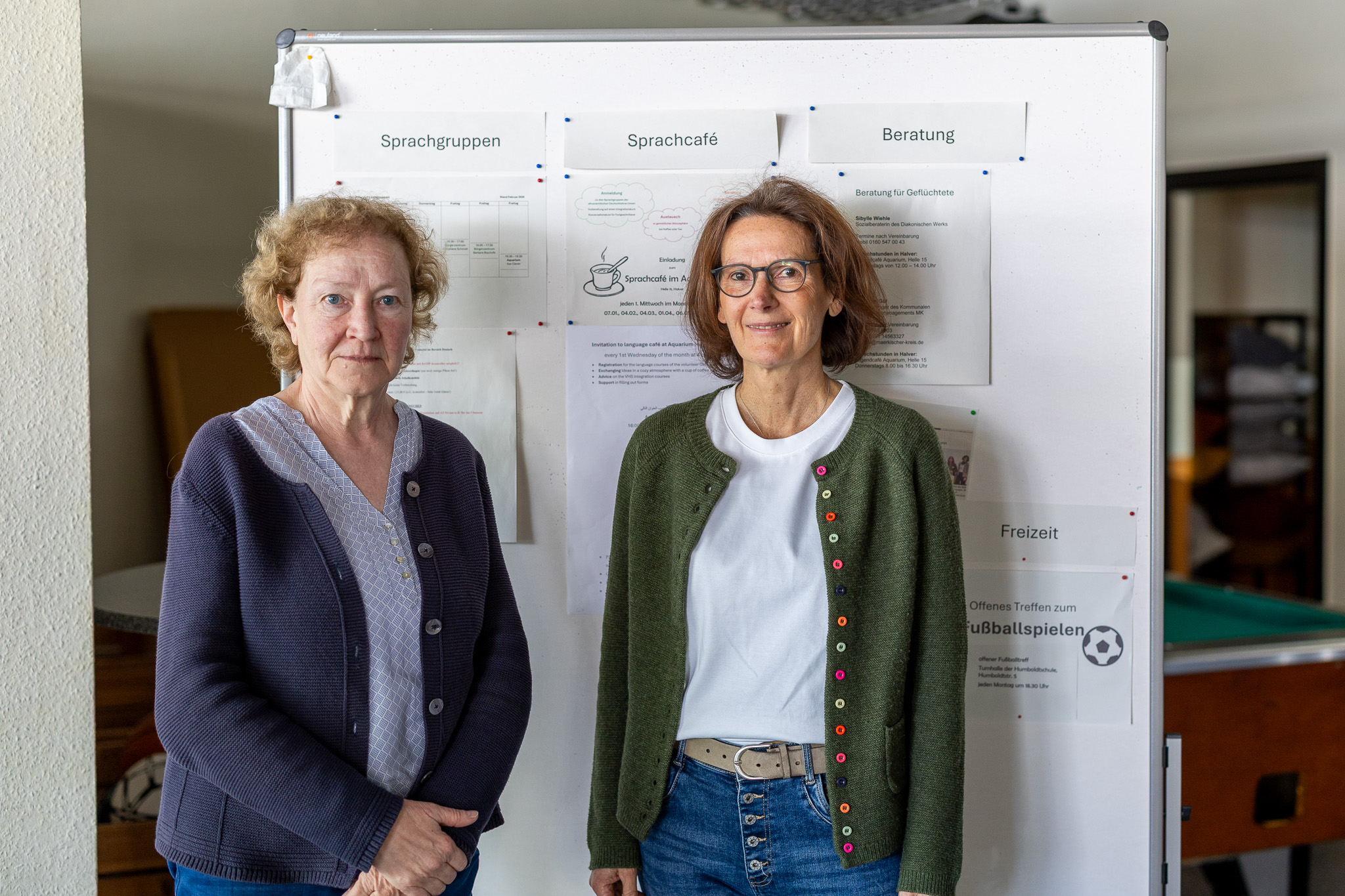 Zwei Frauen mittleren Alters stehen nebeneinander vor Plakaten mit Informationen zu Sprachgruppen, einem Sprachcafé und Beratungsangeboten.
