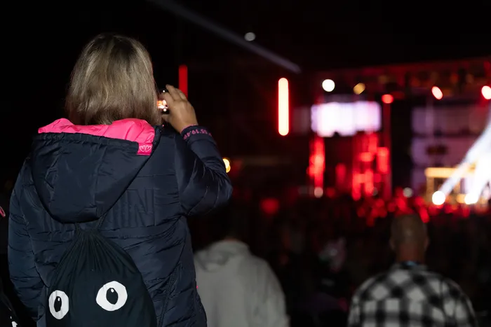 Eine Person mit Rucksack fotografiert mit dem Smartphone eine hell erleuchtete Bühne bei einem Festival bei Nacht.