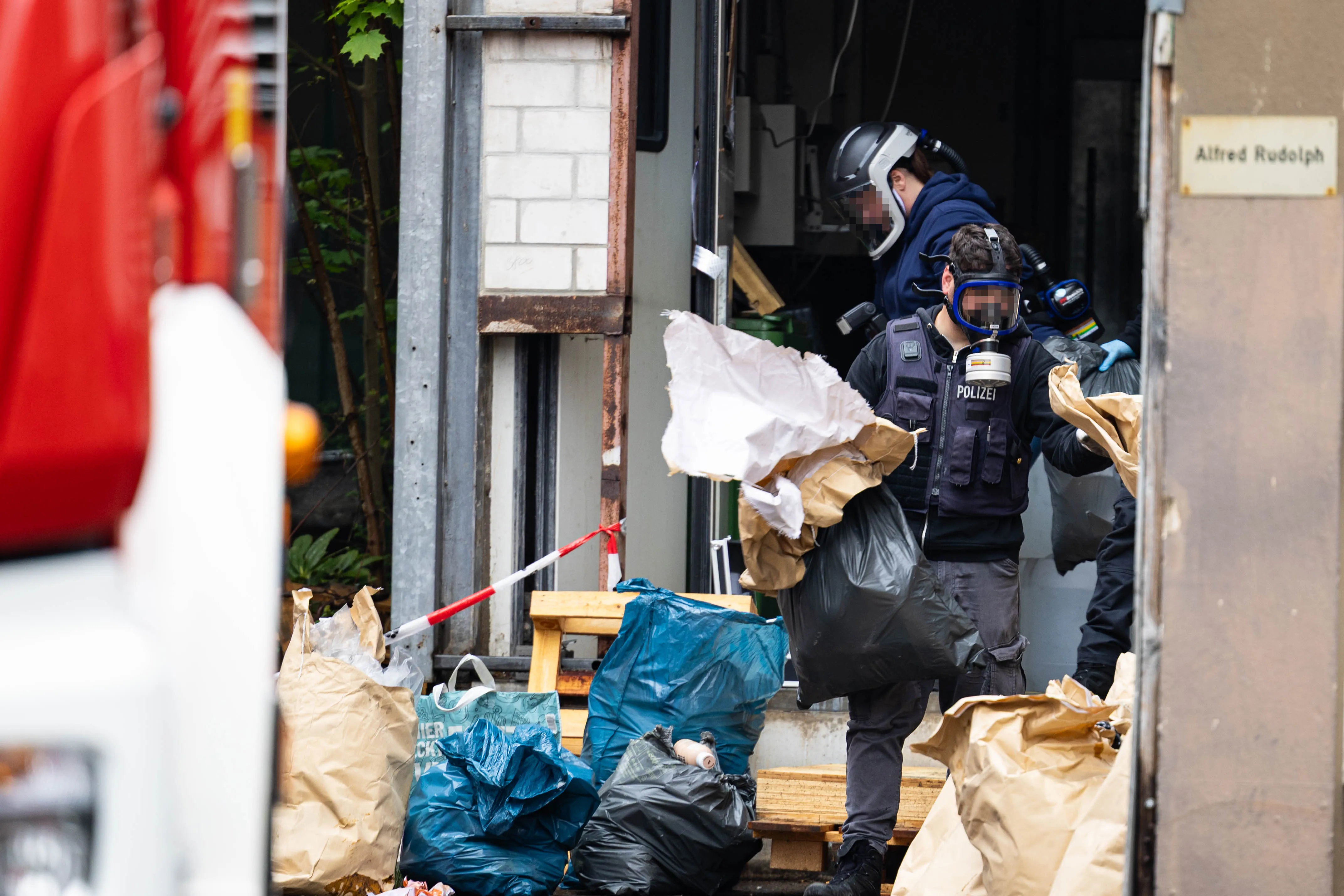 Polizist in Schutzkleidung trägt einen vollen Müllsack aus einem Gebäude, umgeben von Müll und Absperrband. Im Hintergrund ist ein rotes Fahrzeug zu sehen.