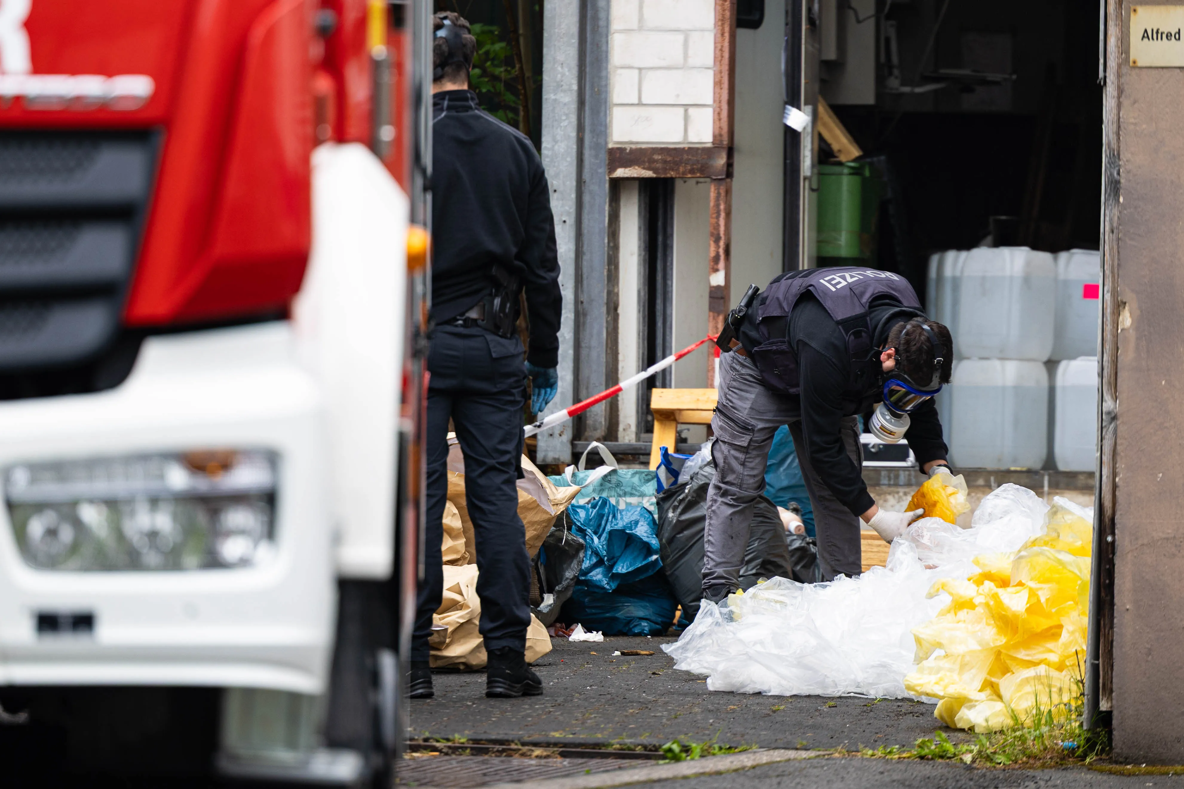 Polizisten in Schutzanzügen untersuchen Müllsäcke und Plastikfolien vor einem Gebäude, während ein roter Lastwagen im Vordergrund steht.