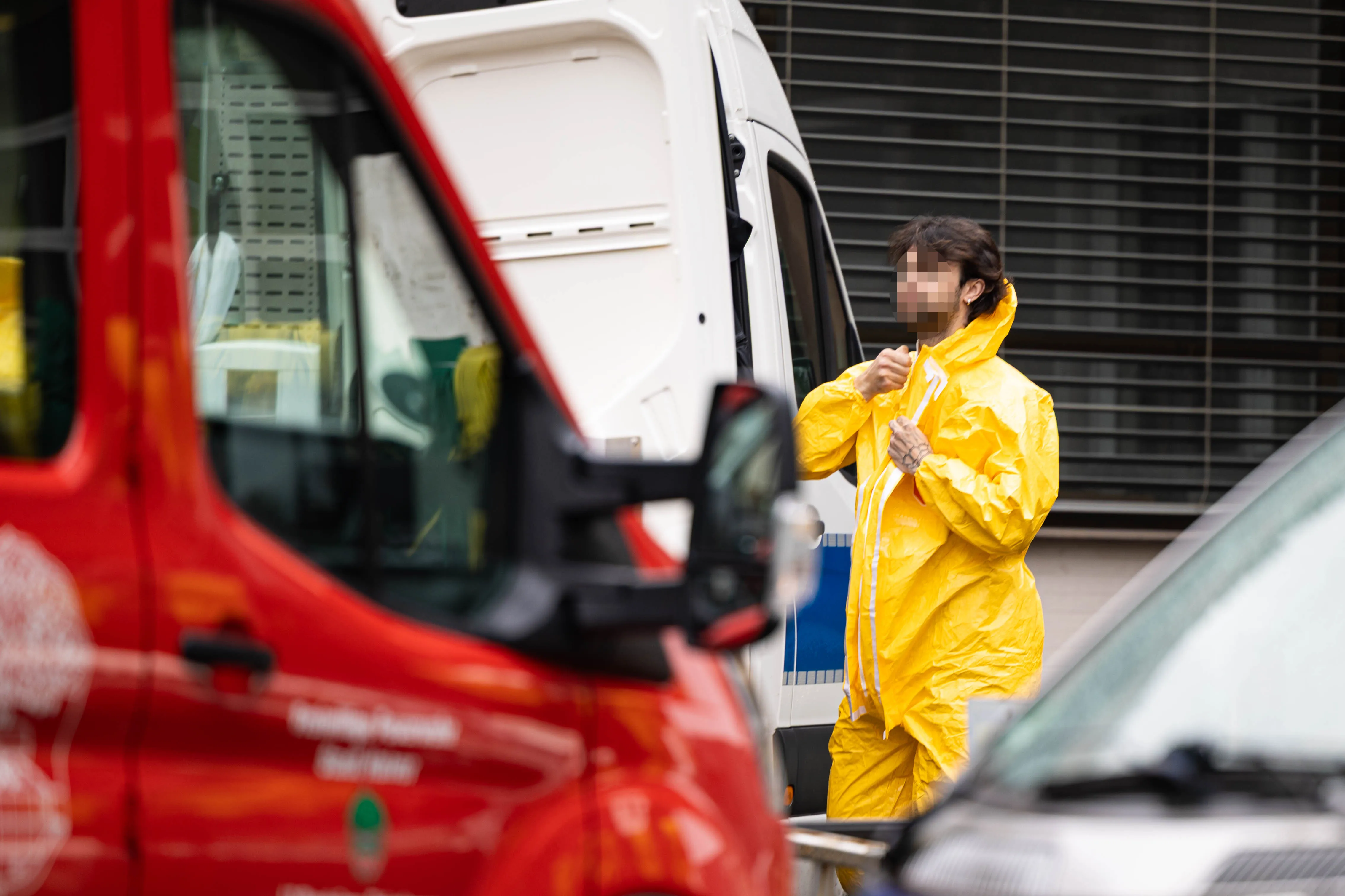 Person in gelbem Schutzanzug steht vor einem Transporter und zieht die Kapuze hoch, während im Hintergrund Einsatzfahrzeuge zu sehen sind.
