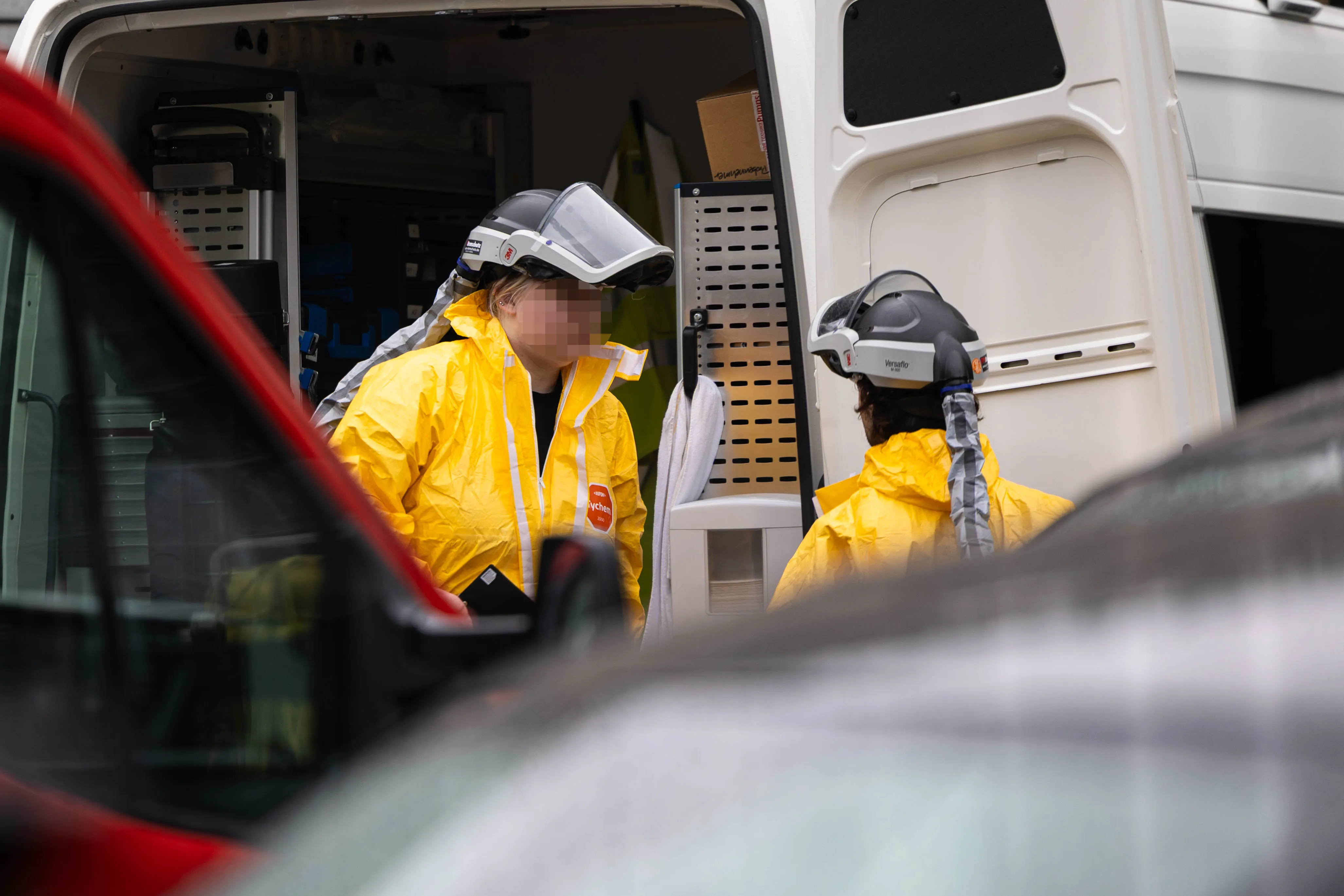 Zwei Personen in gelben Schutzanzügen und mit Atemschutzmasken stehen vor einem Transporter mit geöffneter Hecktür. Im Hintergrund sind weitere Einsatzkräfte und Ausrüstung erkennbar.