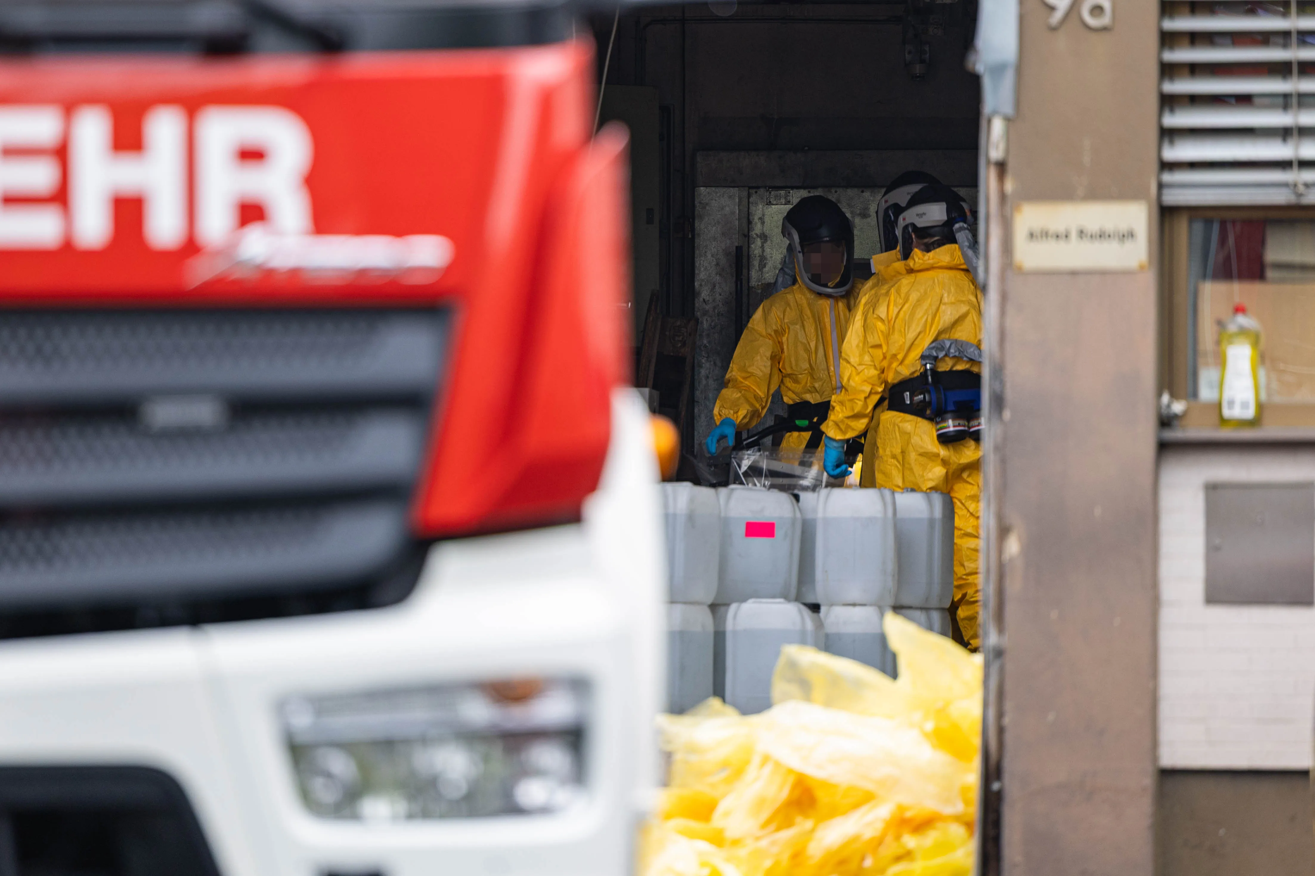 Zwei Personen in gelben Schutzanzügen und Atemschutzmasken arbeiten in einem Transporter, während im Hintergrund ein roter LKW zu sehen ist.