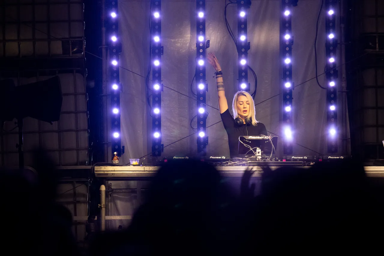 DJane Nusha performt mit erhobenem Arm hinter ihrem Mischpult vor einer Kulisse aus vertikalen blauen Lichtleisten.