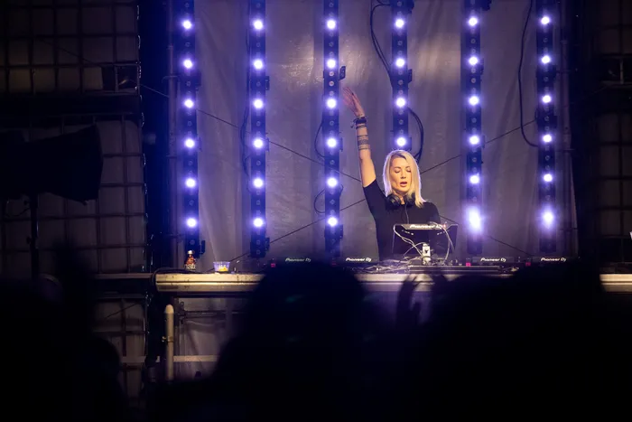 DJane Nusha performt mit erhobenem Arm hinter ihrem Mischpult vor einer Kulisse aus vertikalen blauen Lichtleisten.