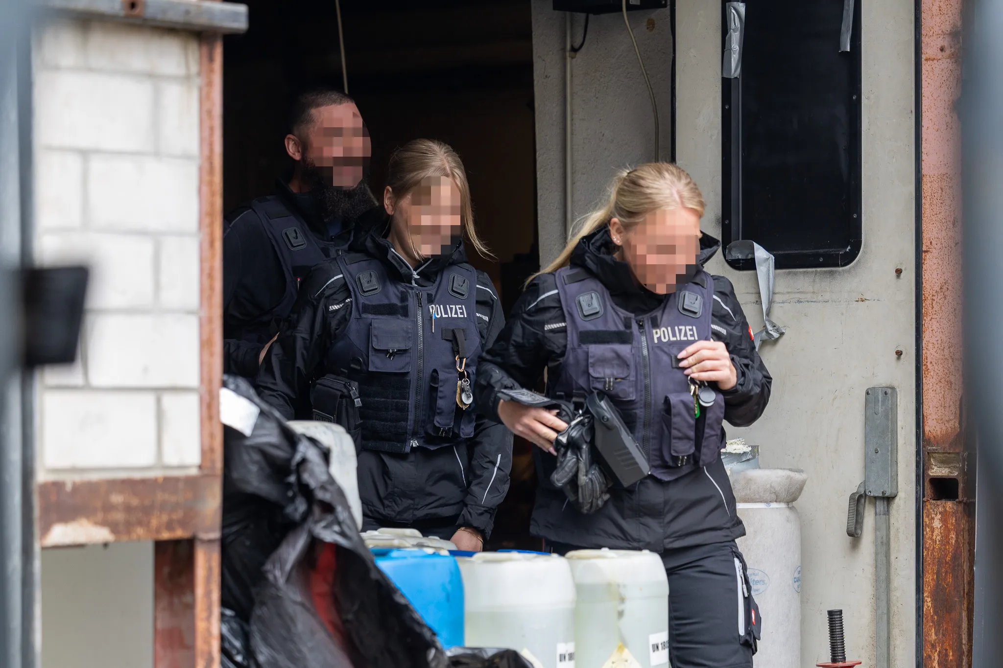 Drei Polizisten in Schutzwesten betreten einen Raum, während ein Entsorgungsdienstleister Kisten und Fässer ausräumt.