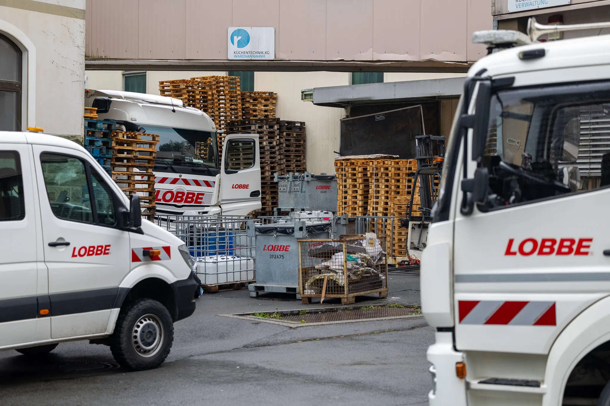 Mehrere Transporter des Entsorgungsdienstleisters Lobbe stehen vor einem Gebäude mit gestapelten Paletten und Containern.