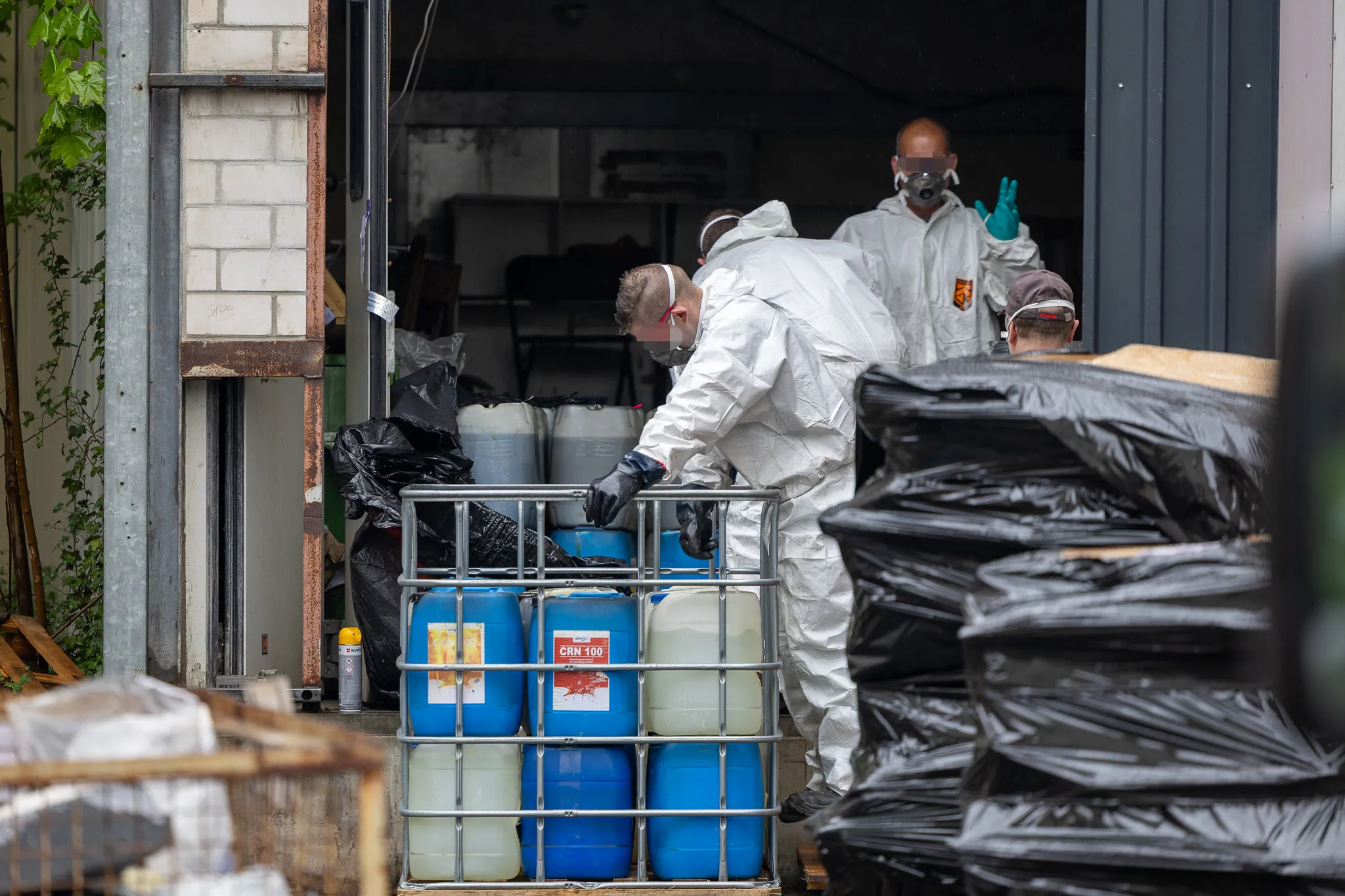 Personen in Schutzanzügen und Masken räumen blaue Fässer und Müllsäcke aus einer Lagerhalle. Im Hintergrund weitere Personen mit Schutzkleidung.