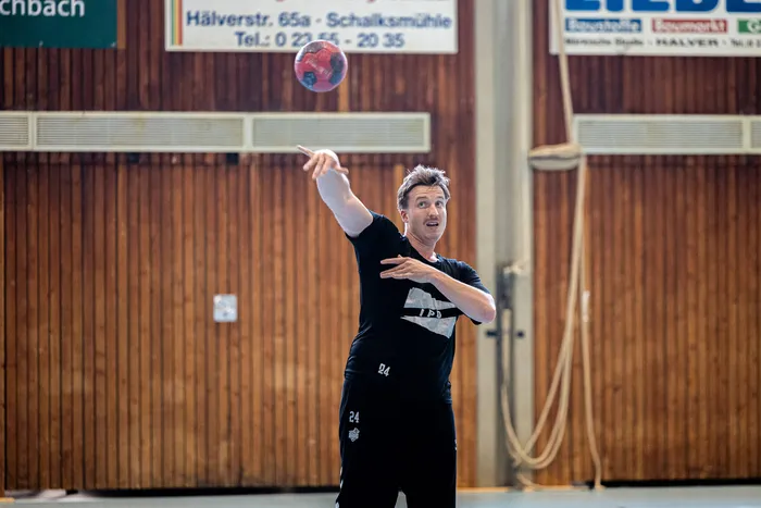 Ein Mann in schwarzer Sportkleidung wirft einen rot gemusterten Handball in einer Sporthalle.