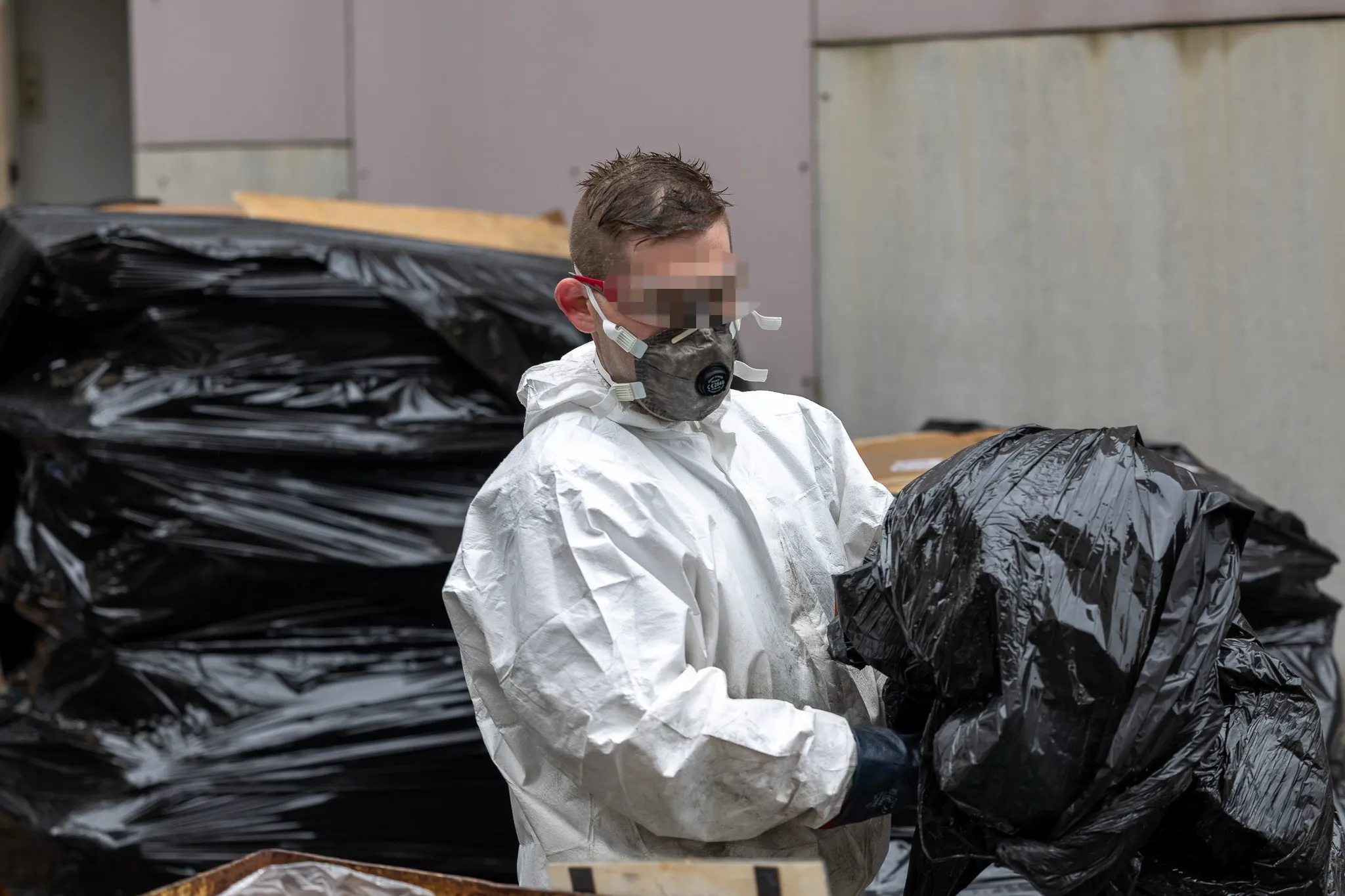 Person in Schutzanzug und Maske hantiert mit schwarzen Müllsäcken vor einem Stapel weiterer Säcke.