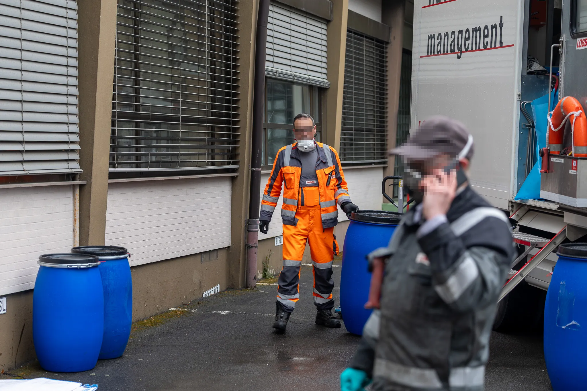Zwei Personen in Schutzanzügen und mit Atemschutzmasken räumen vor einem Gebäude Fässer aus, im Hintergrund ein Fahrzeug mit der Aufschrift „management“.